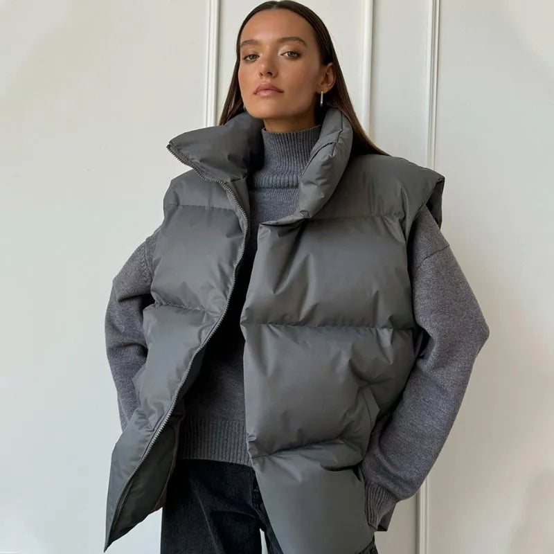 Femme portant une doudoune grise sans manches sur un pull gris, posant devant un mur blanc. Mode hiver, style décontracté, tendance vestimentaire.