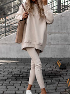 Femme en tenue décontractée beige, portant un sweat oversize et leggings, sac à l'épaule, baskets blanches, sur fond urbain moderne.