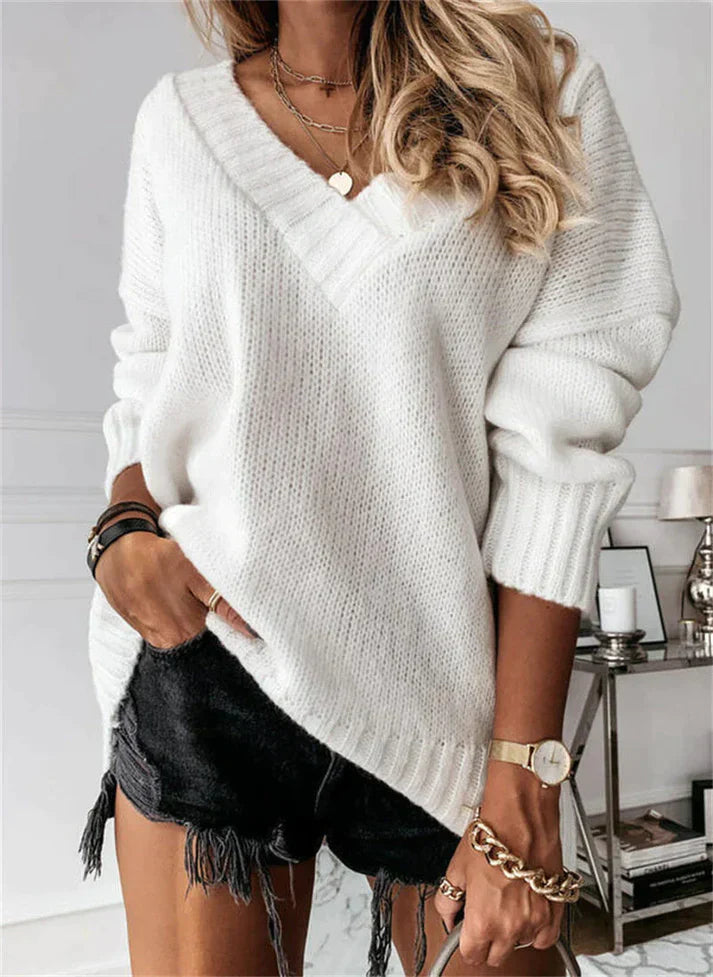 Femme portant un pull blanc en maille avec un col en V, associé à un short noir effiloché. Accessoires dorés et montre élégante. Mode automne chic.