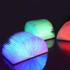 Trois lampes LED en forme de livre, émettant des lumières colorées bleu, vert et rouge, sur une surface réfléchissante sombre.