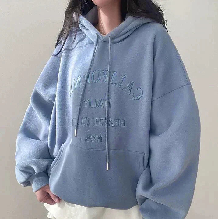 Femme portant un sweat à capuche bleu oversize avec inscription "California" en lettres blanches, style décontracté, mode automne-hiver tendance.