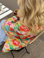 Femme en manteau fleuri coloré, assise sur une chaise en métal, cheveux blonds ondulés, buvant une boisson chaude. Mode printemps, style décontracté.