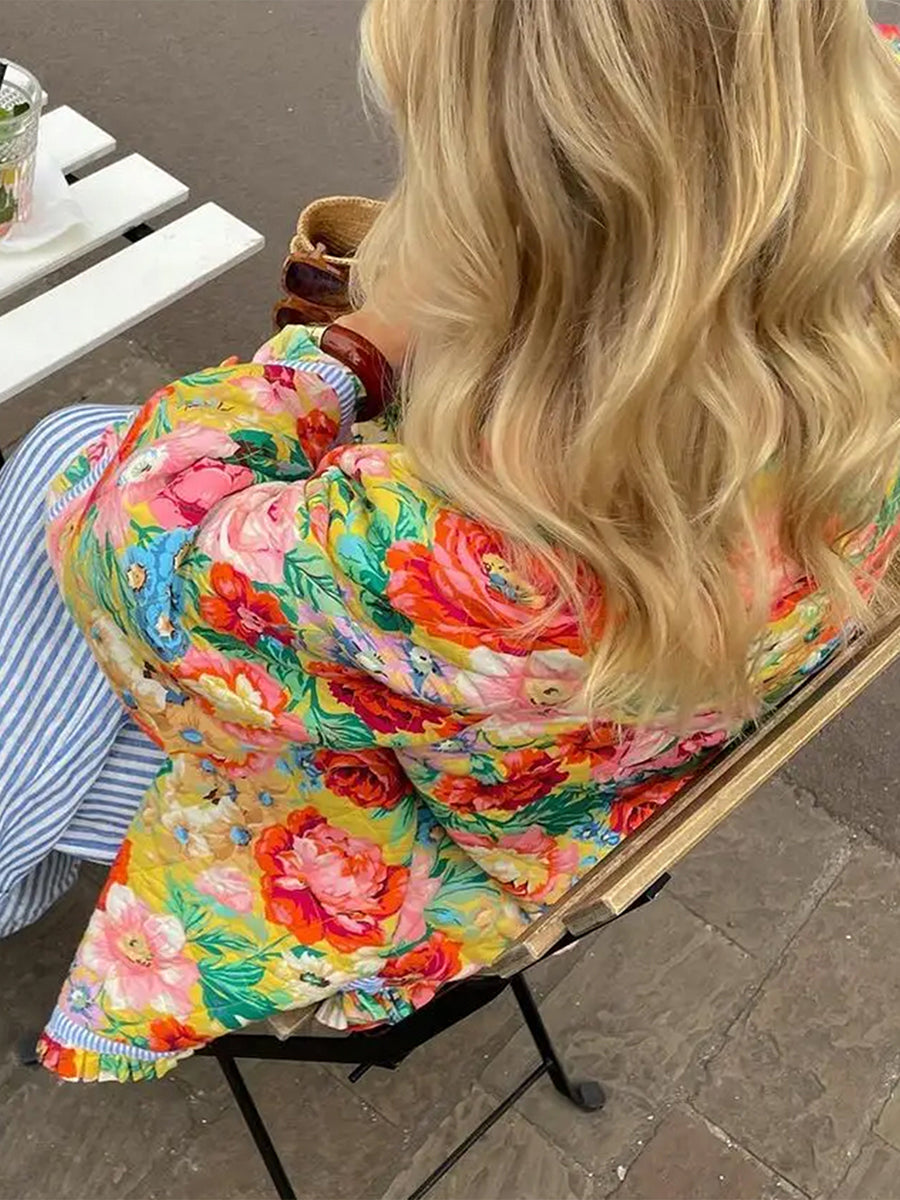 Femme en manteau fleuri coloré, assise sur une chaise en métal, cheveux blonds ondulés, buvant une boisson chaude. Mode printemps, style décontracté.