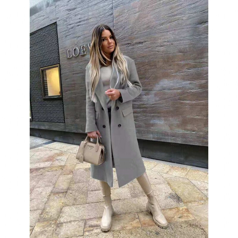Femme élégante portant un manteau gris long, bottes beiges et sac à main beige devant un mur moderne. Mode automne chic, style urbain.