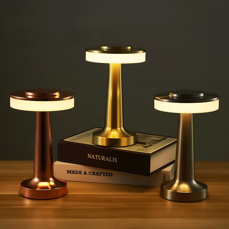 Trois lampes de table LED modernes en métal doré, cuivre et argent, posées sur des livres, éclairant un bureau en bois sombre.