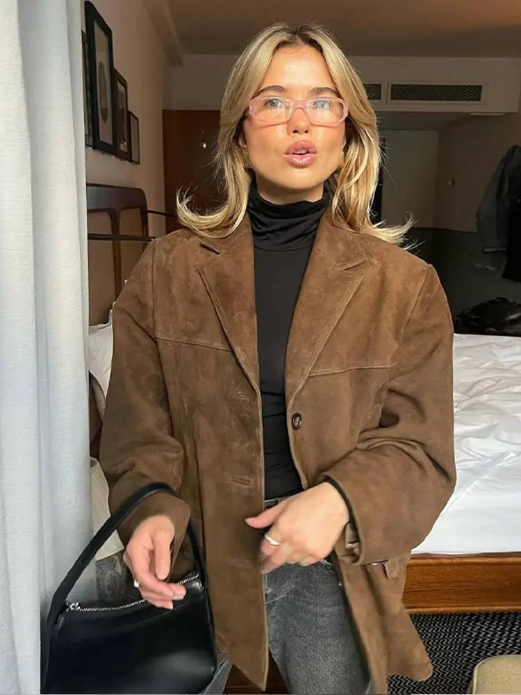 Femme blonde portant une veste en daim marron, col roulé noir et lunettes, tenant un sac à main noir dans une chambre moderne. Mode automne chic.