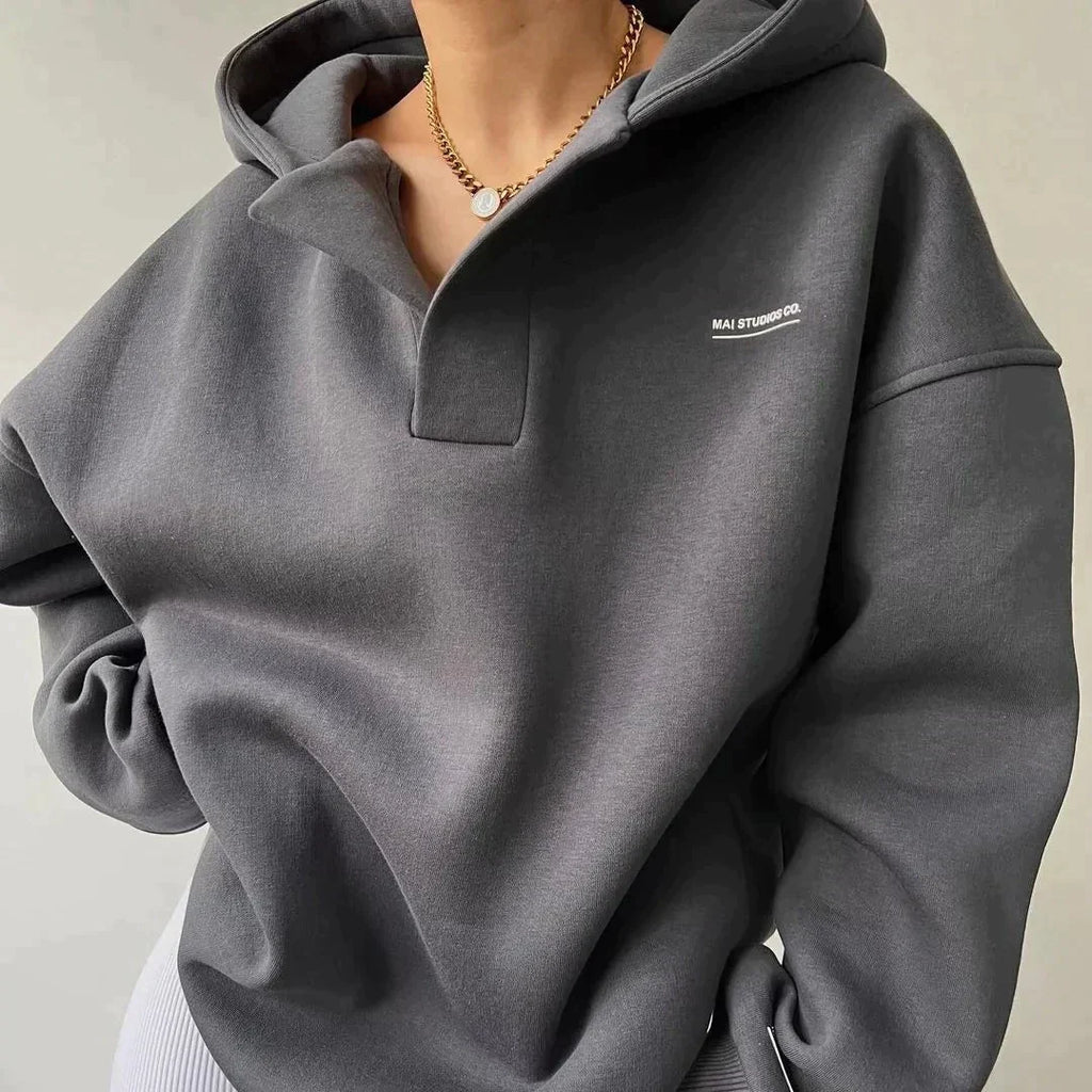 Sweat à capuche gris foncé, col boutonné, porté par une personne, avec collier doré. Mode décontractée, tendance automne-hiver, style urbain.