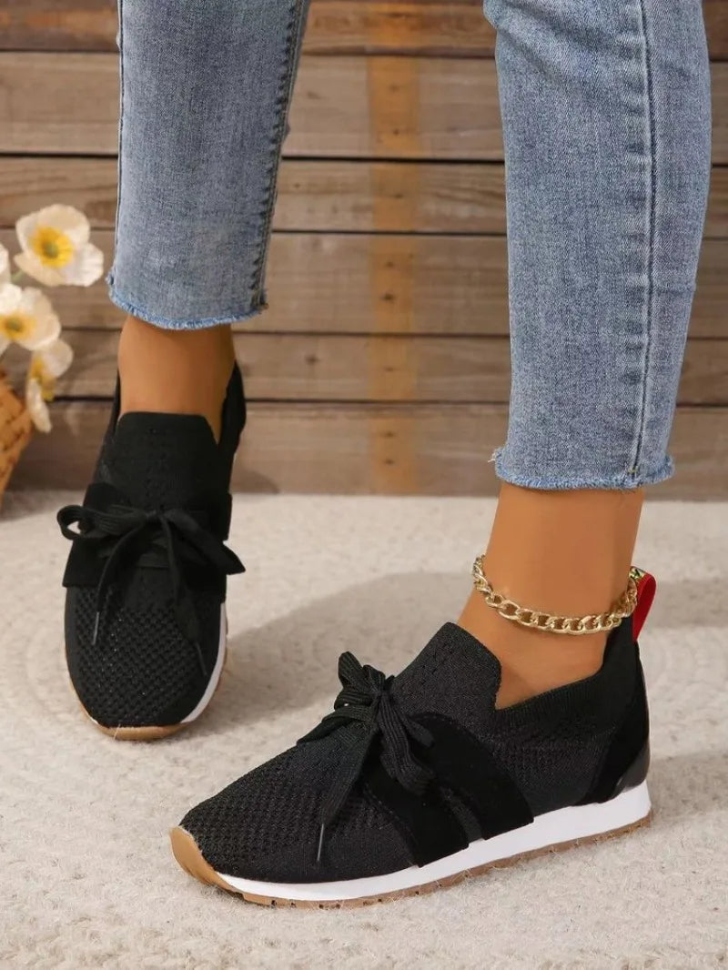 Jeans et baskets noires en maille avec lacets, portées par une personne sur fond de bois. Chaussures décontractées tendance, mode féminine.