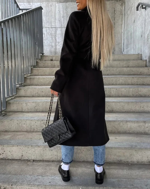 Femme en manteau noir, sac à main en cuir matelassé, jeans, escaliers en béton. Mode automne, style urbain chic, cheveux longs blonds.
