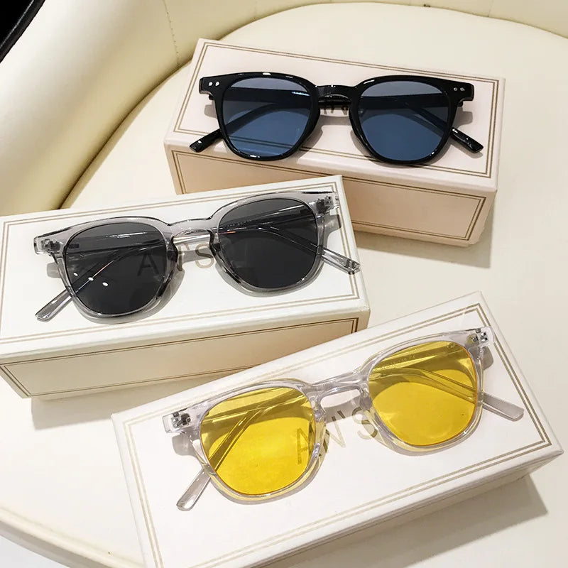 Trois paires de lunettes de soleil élégantes sur des boîtes : noires avec verres bleus, transparentes avec verres gris, et transparentes avec verres jaunes.