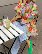 Femme assise en extérieur, portant un manteau fleuri coloré, pantalon rayé bleu et blanc, chaussures vertes, buvant une boisson froide à une table blanche.