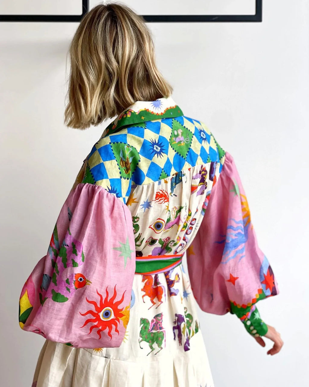 Femme portant une robe colorée avec motifs géométriques et symboles mystiques, manches bouffantes roses, fond blanc. Mode bohème chic, tendance.