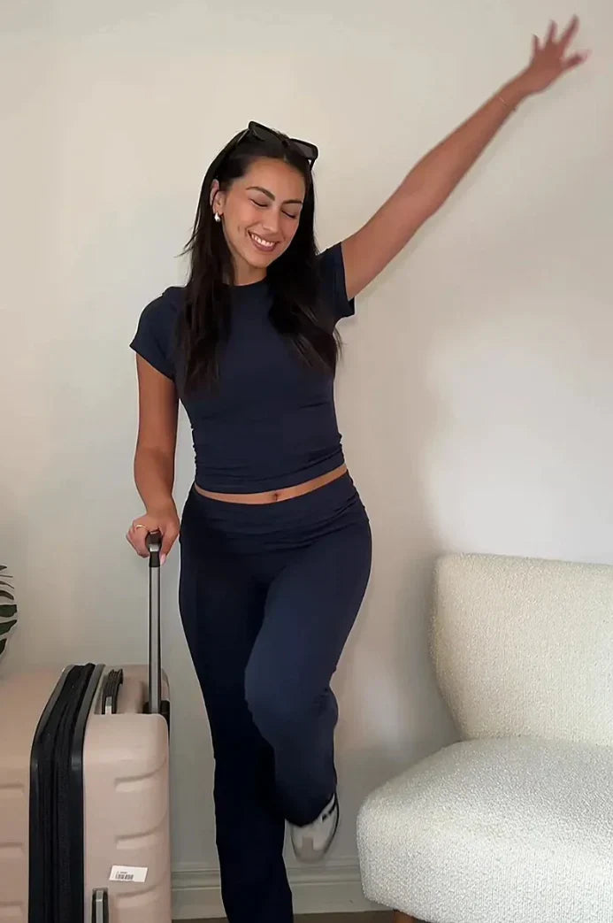 Jeune femme souriante en tenue décontractée bleue, posant avec une valise beige dans un salon moderne. Voyage, mode, détente.