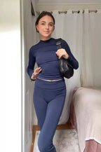 Jeune femme en tenue de sport bleue, posant dans une chambre lumineuse, portant un sac à main noir. Mode fitness, style décontracté, intérieur moderne.