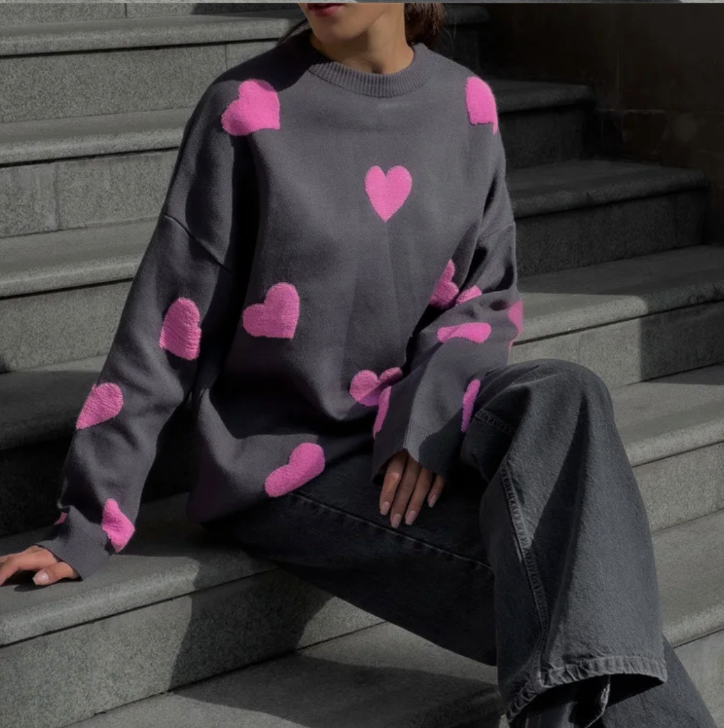 Femme assise sur des marches en pull gris à motifs cœurs roses, pantalon large gris. Mode automne, style décontracté, tendance vêtements cœur.