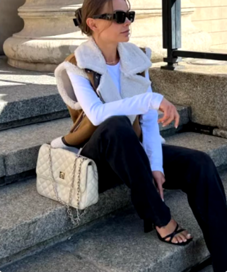 Femme assise sur des marches en pierre, portant des lunettes de soleil, un gilet en fourrure, un sac à main Chanel blanc, et des sandales noires.