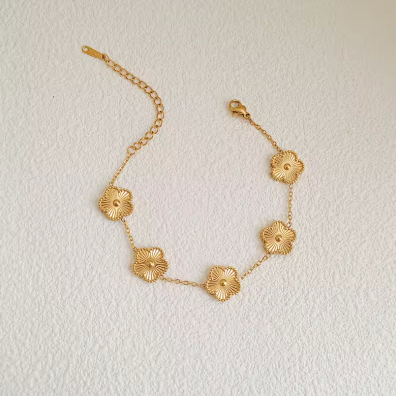 Bracelet en or avec cinq pendentifs en forme de fleur, chaîne délicate et fermoir mousqueton, posé sur une surface texturée. Accessoire élégant.