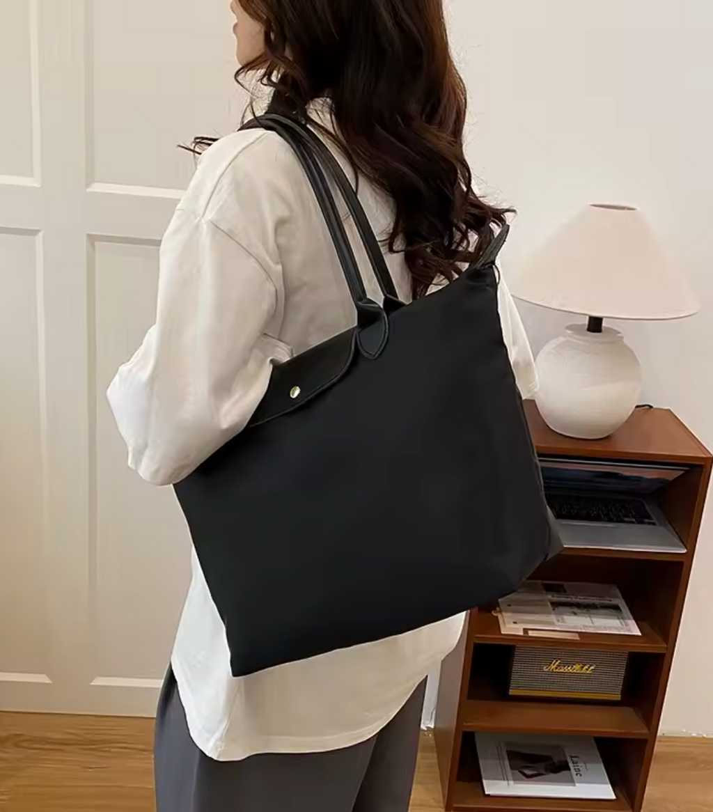 Femme portant un grand sac à main noir en toile, style décontracté, dans un intérieur moderne avec lampe et étagère en bois. Mode féminine élégante.