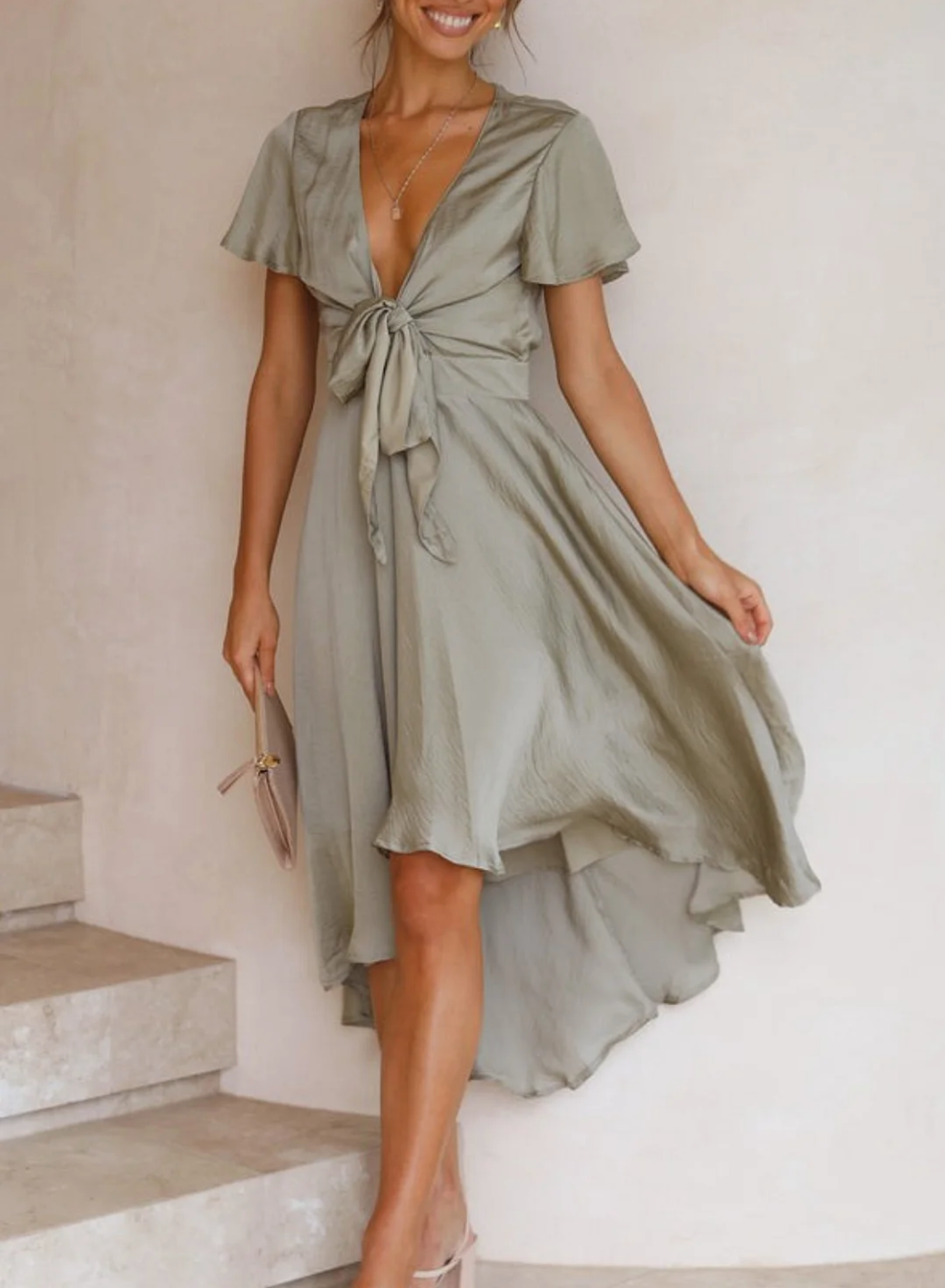 Robe élégante en satin vert olive avec nœud à la taille, manches courtes et ourlet asymétrique, portée par une femme souriante sur des escaliers en pierre.