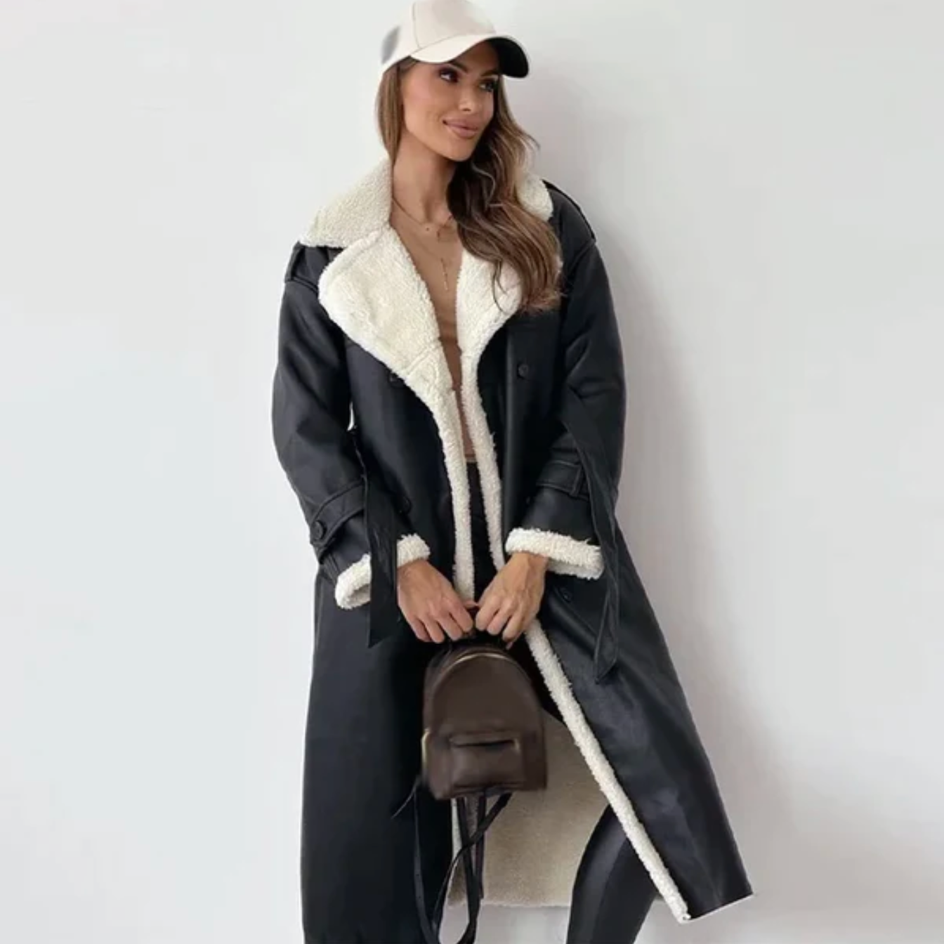 Femme portant un long manteau en cuir noir avec doublure en fourrure blanche, casquette blanche et sac à dos marron. Mode automne-hiver élégante.