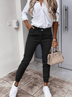 Femme en chemise blanche et pantalon noir, tenant un sac à main beige Prada, portant des baskets blanches, posant devant une porte moderne.