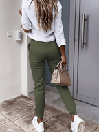 Femme de dos en chemise blanche et pantalon vert, tenant un sac à main beige, devant une porte moderne. Mode décontractée, style urbain.