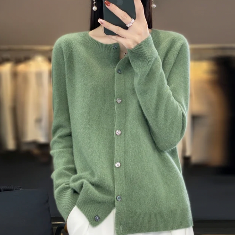 Femme portant un cardigan vert en laine avec boutons, posant devant un fond flou de vêtements suspendus. Mode automne, style élégant et décontracté.