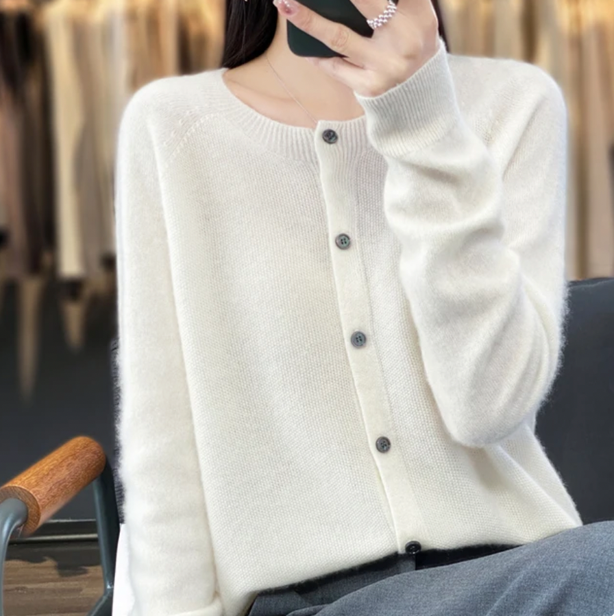 Femme portant un cardigan en laine blanc, assise sur une chaise, tenant un smartphone. Mode automne-hiver, style élégant et décontracté.