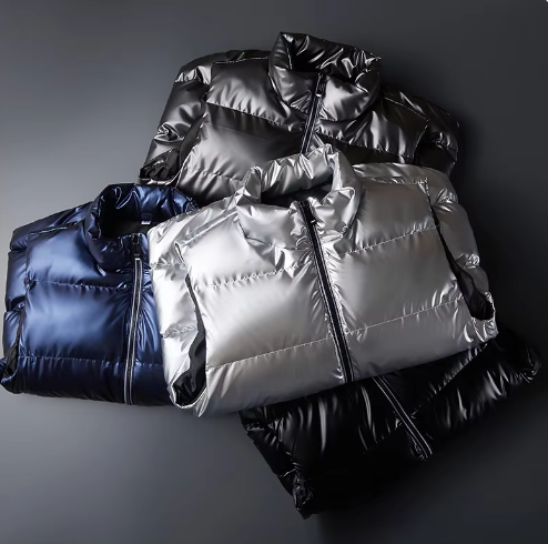 Quatre doudounes en tissu brillant, couleurs noir, argent et bleu, posées sur une surface grise. Mode hiver, vêtements chauds, style moderne.