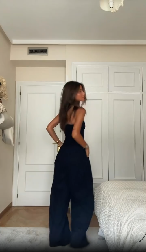 Femme portant une combinaison noire élégante dans une chambre moderne, posant devant une armoire blanche. Mode féminine, tenue chic, style contemporain.