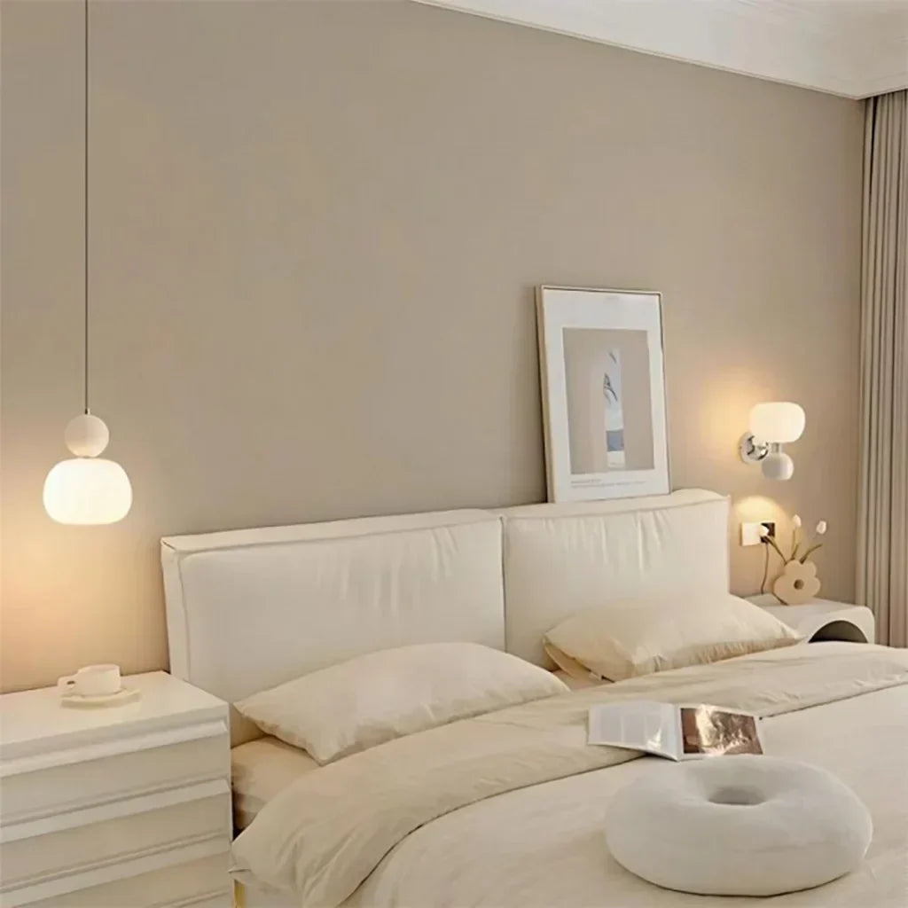 Chambre moderne avec lit double beige, lampes suspendues, tableau encadré, table de chevet blanche et coussin rond. Décoration intérieure élégante.