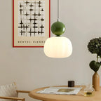 Suspension moderne avec abat-jour blanc et vert, éclairant une table en bois. Décor minimaliste avec affiche artistique et plante en pot.