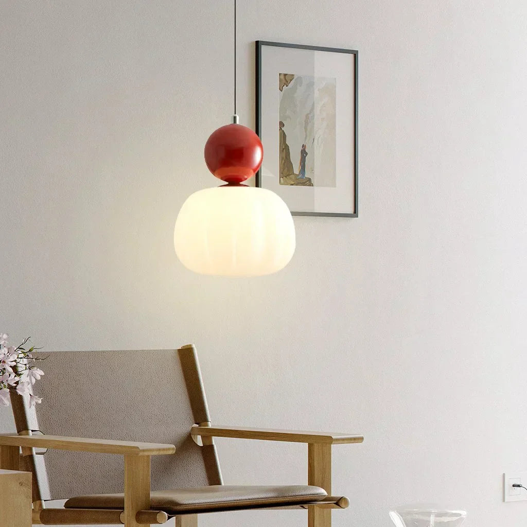 Suspension moderne avec abat-jour blanc et détail rouge, éclairant un coin salon minimaliste avec fauteuil en bois et tableau encadré.
