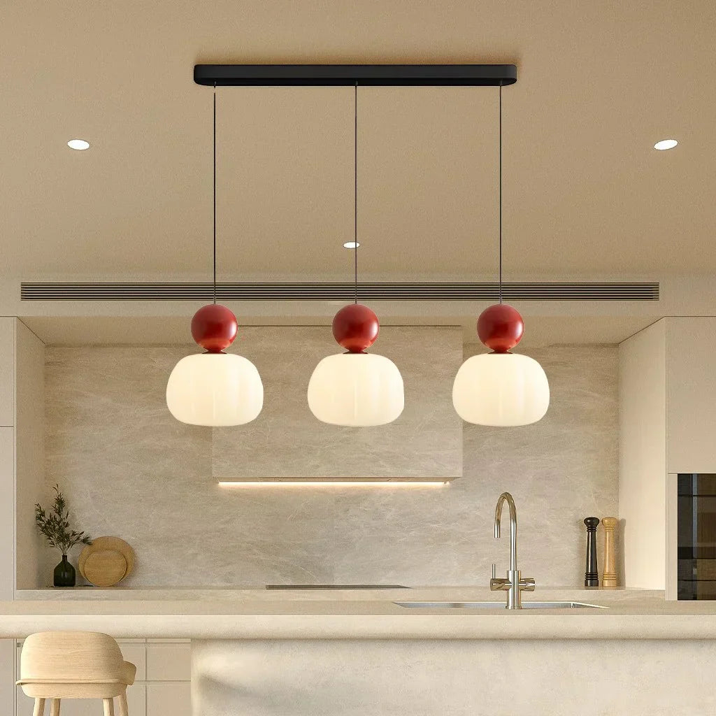 Suspension moderne à trois lampes avec abat-jours blancs et détails rouges, installée au-dessus d'un îlot de cuisine minimaliste en marbre beige.