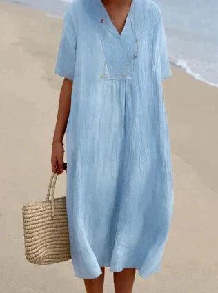 Robe d'été bleue en lin, manches courtes, portée sur une plage. Sac en osier à la main. Mode plage, tenue estivale élégante et décontractée.