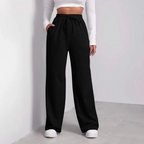 Femme portant un pantalon noir ample taille haute, avec un haut blanc à manches longues, debout sur un sol gris. Mode décontractée élégante.