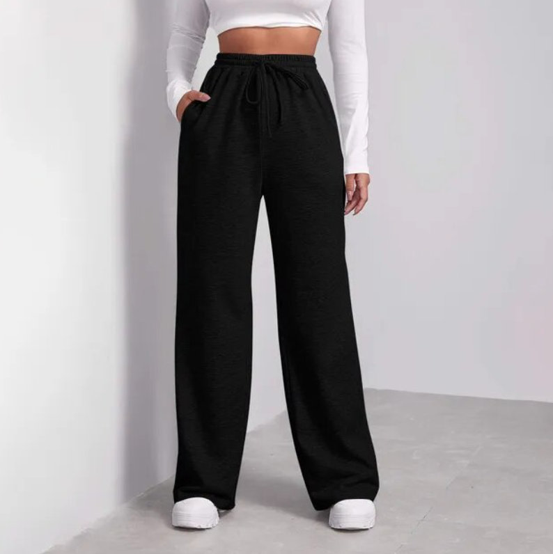 Femme portant un pantalon noir ample avec un haut blanc à manches longues, posant dans un intérieur moderne. Mode décontractée, style tendance.