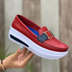 Chaussure mocassin rouge en cuir avec semelle épaisse blanche, boucle décorative dorée et détails bleus, tenue par une main avec ongles peints.