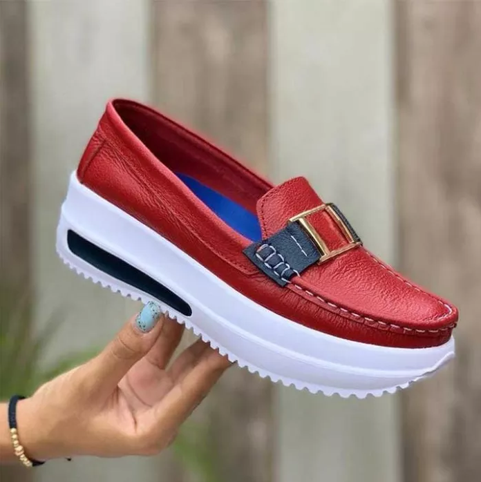 Chaussure mocassin rouge en cuir avec semelle épaisse blanche, boucle décorative dorée et détails bleus, tenue par une main avec ongles peints.