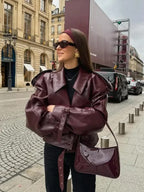 Femme portant une veste en cuir bordeaux, lunettes de soleil et sac assorti, posant dans une rue parisienne. Mode automne, style urbain chic.