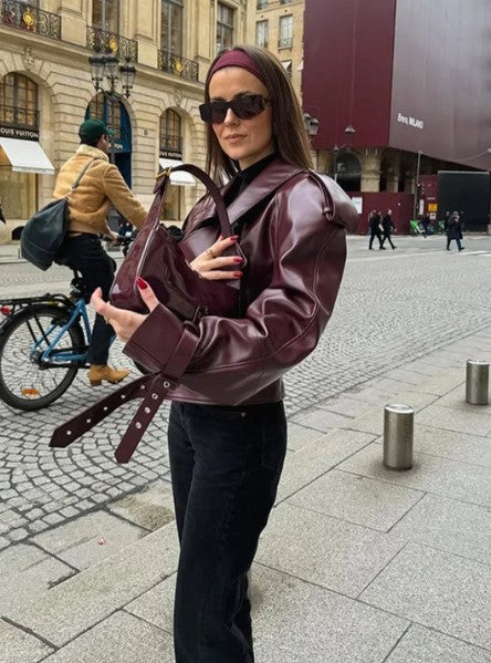 Femme portant une veste en cuir bordeaux et lunettes de soleil, tenant un sac assorti, dans une rue parisienne animée. Mode urbaine chic.