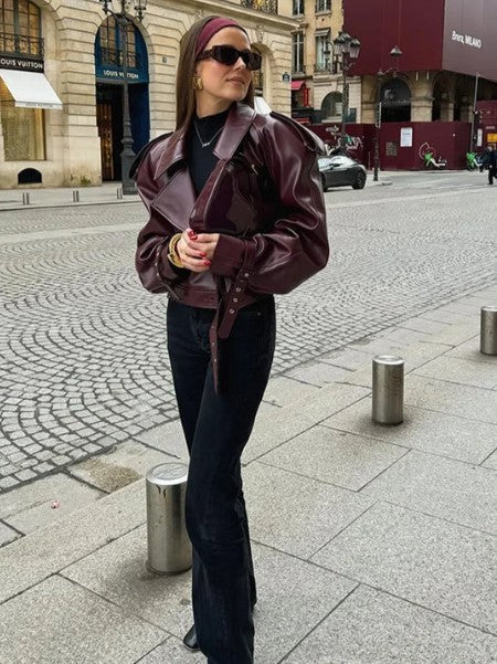 Femme portant une veste en cuir bordeaux, lunettes de soleil et bandeau, posant dans une rue pavée parisienne. Mode urbaine chic, style automne.