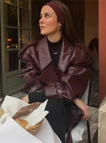 Femme portant un manteau en cuir marron, assise à une table de café avec des viennoiseries, ambiance élégante et moderne.