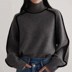 Femme portant un pull gris à col roulé avec bordures noires, manches longues, associé à un jean bleu. Mode automne-hiver, style décontracté.