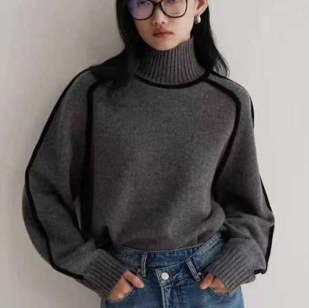 Jeune femme portant un pull gris à col roulé et lunettes, posant contre un mur. Mode automne-hiver, style décontracté, tendance féminine.