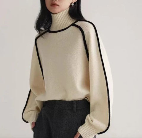 Femme portant un pull col roulé beige avec bordures noires, manches longues amples, associé à un pantalon gris. Mode automne-hiver élégante.