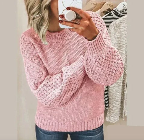 Femme portant un pull rose en tricot texturé, manches longues, tenant un smartphone devant un miroir. Mode automne-hiver, style décontracté.