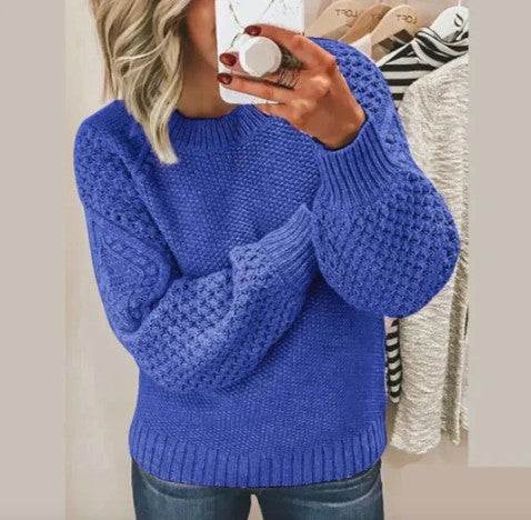 Femme portant un pull bleu en tricot texturé, manches longues, tenant un smartphone. Mode automne-hiver, style décontracté, tendance féminine.