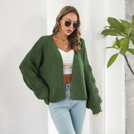 Femme portant un cardigan vert en maille, lunettes de soleil, top blanc et jean bleu, posant près d'une plante d'intérieur. Mode automne chic.