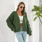 Femme portant un cardigan vert en maille, lunettes de soleil, top blanc et jean bleu, posant près d'une plante d'intérieur. Mode automne chic.